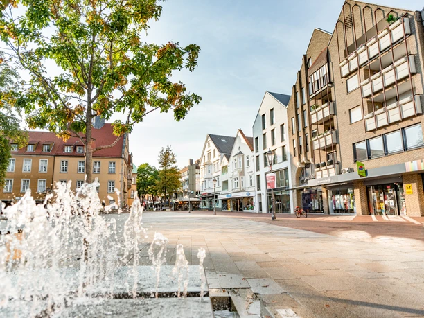 Belebter Platz mit Wasserspielen, umgeben von modernen Gebäuden und Bäumen unter blauem Himmel.