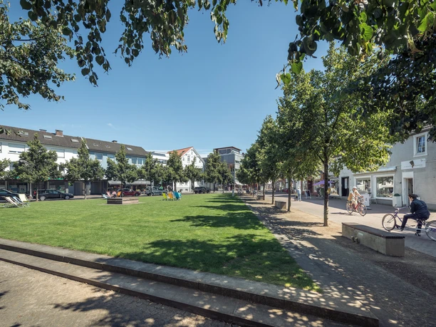 Grüne Parkanlage mit Bäumen, Radfahrer und Fußgänger bei sonnigem Wetter, umgeben von Gebäuden.