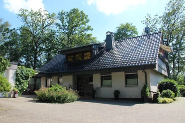 049_Hausfront (1).JPG Ein einstöckiges Haus mit grau-gedecktem Dach, steinverkleideten Wänden und üppigem Grün im Hintergrund.