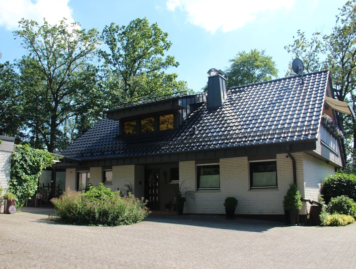 049_Hausfront (1).JPG Ein einstöckiges Haus mit grau-gedecktem Dach, steinverkleideten Wänden und üppigem Grün im Hintergrund.