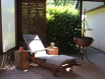 053_Pergola1.jpg Sonnenlicht fällt auf eine Holzliege mit Polster auf einer Terrasse, umgeben von grünen Büschen.