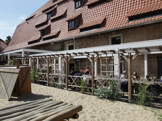 Der Außenbereich des Alten Brauhauses zu Fallersleben zeigt einen gemütlichen Biergarten mit Holztischen unter einer Pergola. Links ist ein kleiner Holzspielplatz auf sandigem Boden zu sehen. Der Backsteinbau im Hintergrund trägt ein rotes Ziegeldach mit Gauben.