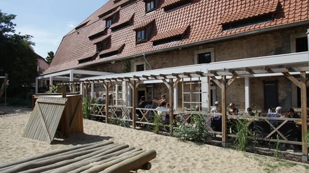 Außenbereich vom Alten Brauhaus zu Fallersleben Der Außenbereich des Alten Brauhauses zu Fallersleben zeigt einen gemütlichen Biergarten mit Holztischen unter einer Pergola. Links ist ein kleiner Holzspielplatz auf sandigem Boden zu sehen. Der Backsteinbau im Hintergrund trägt ein rotes Ziegeldach mit Gauben.
