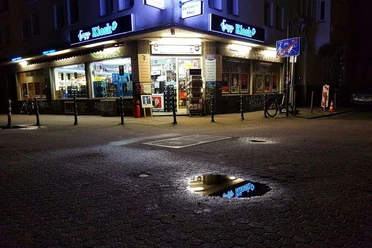 Jazz Kiosk Der Jazz-Kiosk bei Nacht zeigt ein beleuchtetes Geschäft in urbaner Umgebung mit reflektierendem Schaufenster und Regenpfütze.