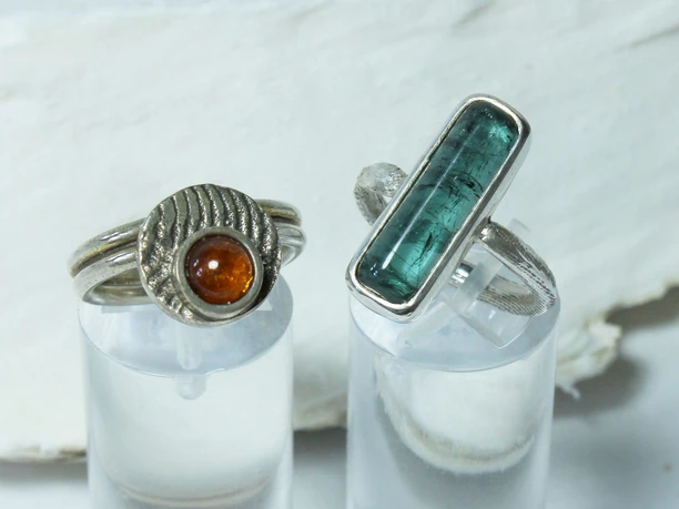 A gauche, une bague avec un disque rond et une pierre précieuse orange et, à droite, une bague avec une pierre précieuse bleue allongée de Sarah Gaus, produits régionaux