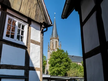 Die Ansicht zeigt mittelalterliche Fachwerkhäuser und einen Kirchturm, eingerahmt von zwei Häuserdächern.