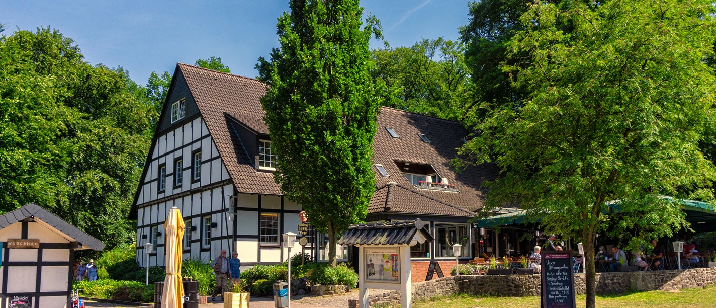 Café-Restaurant am Ausflugsziel Rubbenbruchsee Half-timbered house with café terrace on the edge of the forest, sunny weather, visitors enjoy the outdoor area.