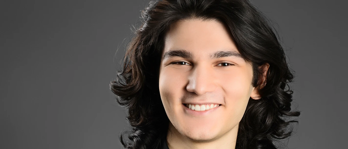 Weltklassik: Kaan Baysal Lächelndes Porträt des Pianisten Kaan Baysal mit dunklem Haar vor grauem Hintergrund.