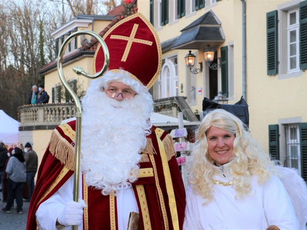 Weihnachtsmarkt Schloss Eulenbroich Sankt Nikolaus mit Bischofsstab und Engel auf einem festlichen Weihnachtsmarkt, im Hintergrund Fachwerkhäuser.