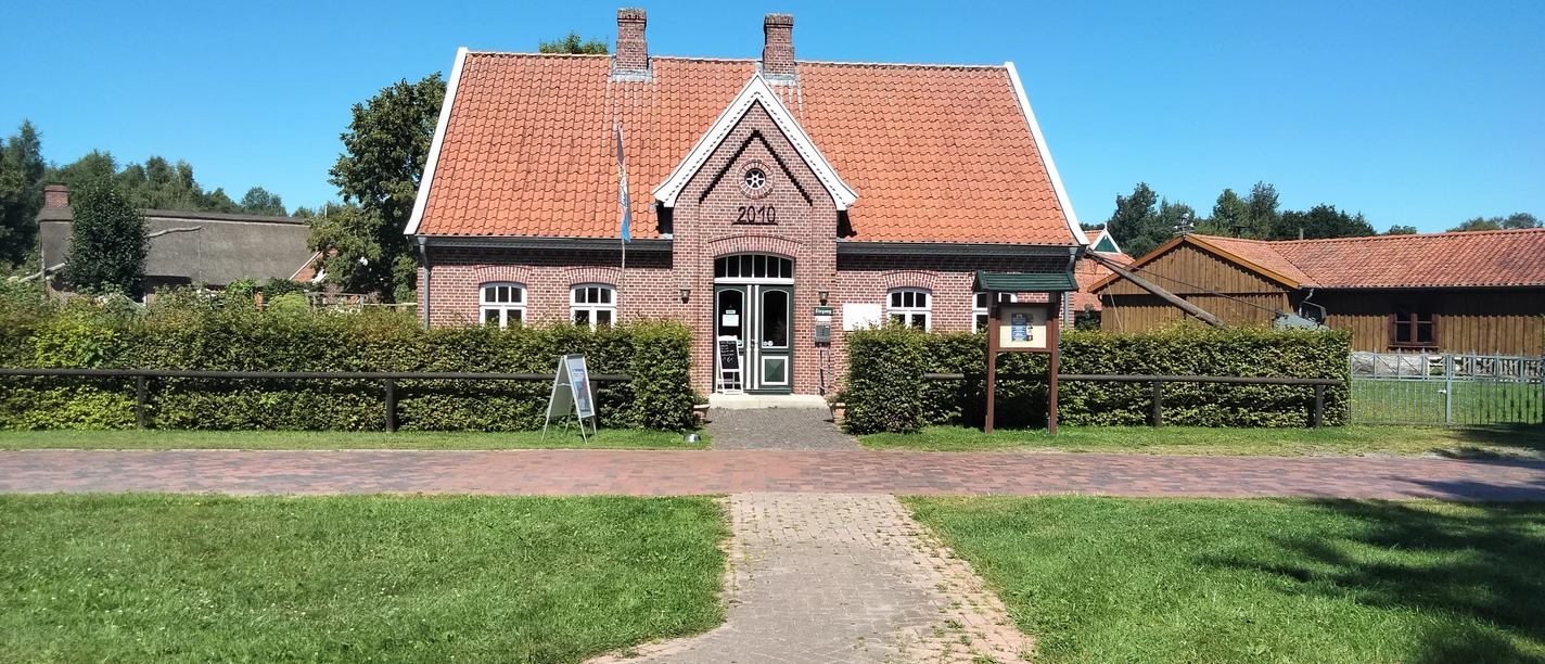 IMG20220811124054.jpg Das Bild zeigt ein charmantes Backsteinhaus mit rotem Ziegeldach inmitten einer grünen Wiese unter einem strahlend blauen Himmel. Vor dem Haus sind gepflegte Büsche und ein gepflasterter Weg sichtbar, der zur Eingangstür führt.