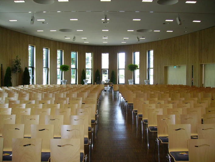 Holzverkleideter Saal mit Reihen aus Stühlen, bodentiefen Fenstern und Pflanzen an den Wänden.