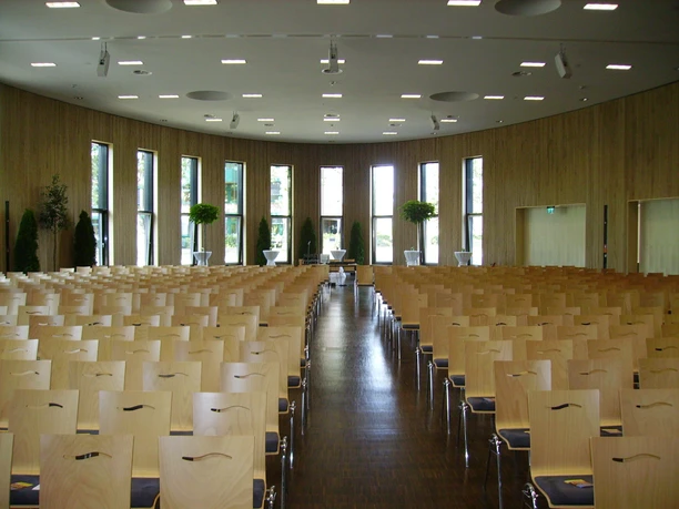 Aula_SHS_CC-BY-SA_Teutoburger_Wald_Stadt_Schloss_Holte-Stukenbrock (2).jpg Holzverkleideter Saal mit Reihen aus Stühlen, bodentiefen Fenstern und Pflanzen an den Wänden.
