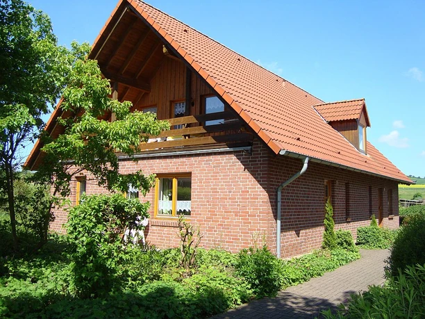 Ferienhaus Backsteinhaus mit rotem Satteldach, umgeben von grünen Sträuchern und Bäumen unter blauem Himmel.