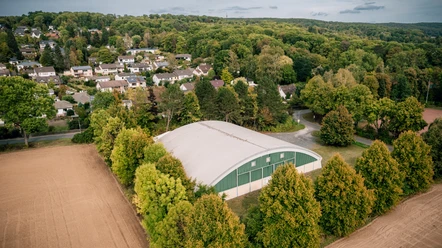 Tennishalle in Einbeck Große Tennishalle mit gewölbtem Dach, umgeben von Bäumen und Wohnhäusern am Waldrand in Einbeck.