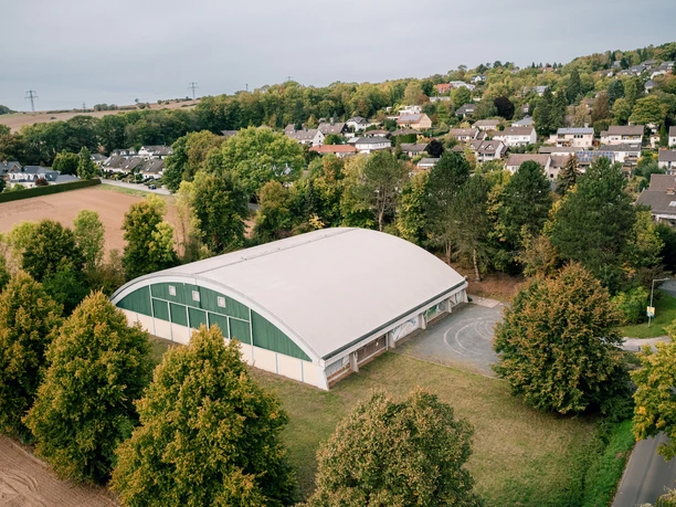 Tennishalle in Einbeck Große Tennishalle mit gewölbtem Dach, umgeben von Bäumen und Wohnhäusern am Rand von Einbeck.