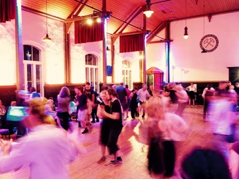 lindy-hop-3.jpg Tanzende Menschen füllen einen festlich beleuchteten Veranstaltungsraum mit hohen Fenstern.
