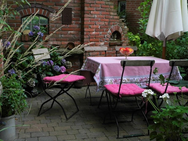 Ein idyllischer Platz zum Entspannen mit vier Klappstühlen um einen Tisch mit Rosa Tischdecke und auf dem Tisch ein höhergesetzte Glaseschale mit Blumen drin liegt , herrlich umgeben von üppigem Grün.
