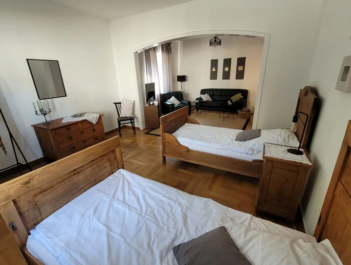 6.2 Wohnraum+Schlafraum.JPG Zwei Einzelbetten aus Holz in einem geräumigen Zimmer, kombiniert mit einem angrenzenden Wohnbereich.