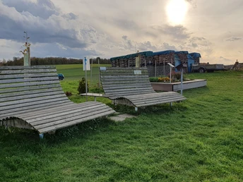 Landschaftsliegen Zwei große Holzliegen stehen auf einer grünen Wiese mit Blick auf Felder, unter einer bewölkten Sonne.