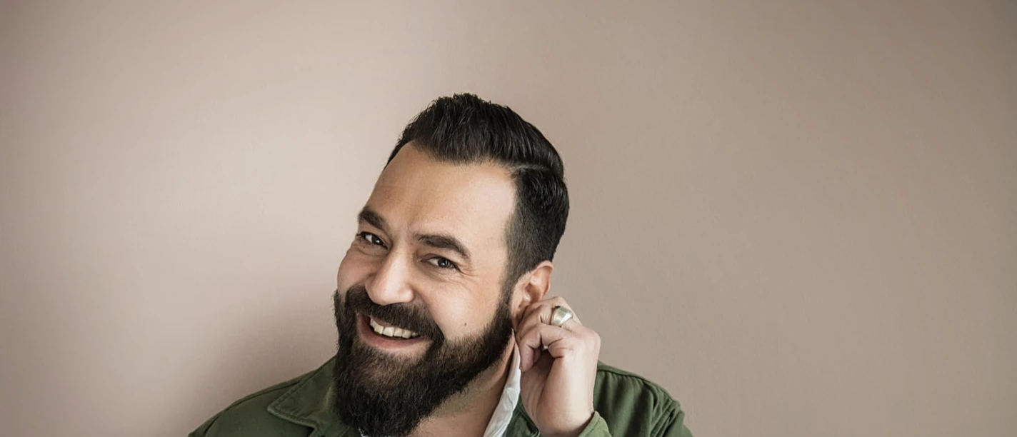 20230825_Laith_Al-Deen_(c)_Chris_Gonz.jpg Singer Laith Al-Deen smiles warmly, wearing a jacket and shirt, standing in front of a neutral background.