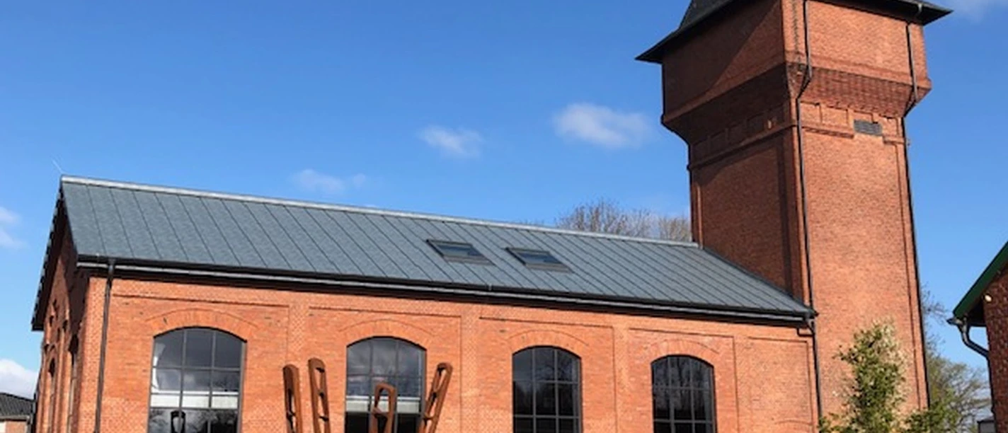 Außenbereich Eisenhütte Das Bild zeigt ein historisches, rotes Backsteingebäude. Es hat große Fenster und einen markanten, spitzen Turm. Der Himmel ist klar und blau.