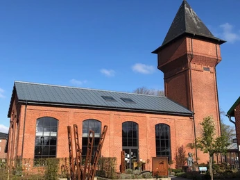 Außenbereich Eisenhütte Das Bild zeigt ein historisches, rotes Backsteingebäude. Es hat große Fenster und einen markanten, spitzen Turm. Der Himmel ist klar und blau.