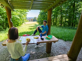 Zwei Personen sitzen an einem Holztisch unter einem hölzernen Pavillon im sommerlichen Wald.