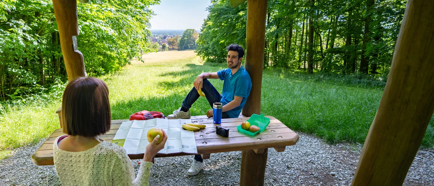 Halle-Hörstation-Teutoburger-Wald-Tourismus-Patrick-Gawandtka-090_lowres.jpg Zwei Personen sitzen an einem Holztisch unter einem hölzernen Pavillon im sommerlichen Wald.