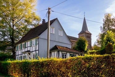 Kirche Delling und Restaurant Fachwerkhaus mit grünen Fensterläden, im Hintergrund ein Kirchturm und blauer Himmel, umgeben von Bäumen.