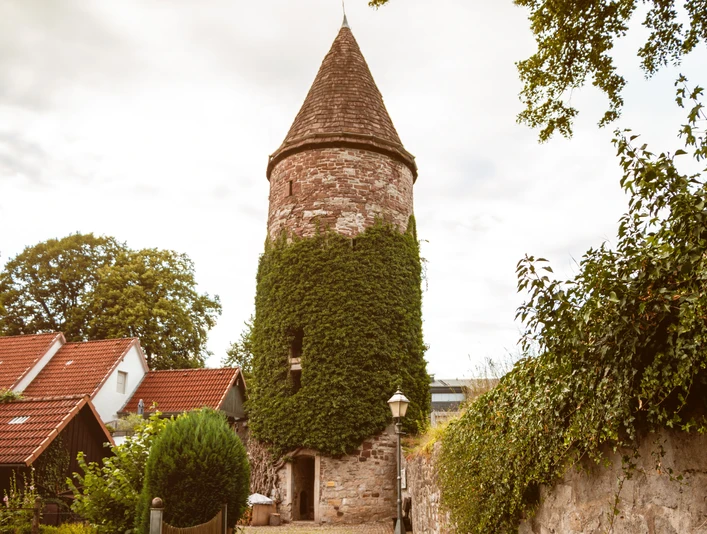 Wehrturm mit runder, begrünten Fassade und spitzem Dach, umgeben von Häusern und Bäumen.