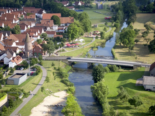 Emmerauenpark aus der Vogelperspektive Vogelperspektive auf einen Park mit Fluss, Brücke und Sitzbereichen, umgeben von grüner Landschaft.