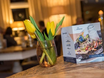 Bad Driburger Gourmet-Tour Gelbe Tulpen in einem kleinen Glasgefäß auf einem Holztisch, im Hintergrund eine Menükarte der Gourmet-Tour.