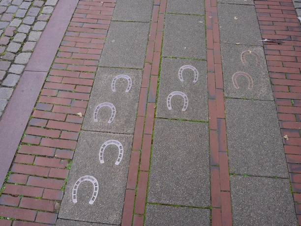 Pflasterweg mit Hufeisenmustern im Bodenbelag, flankiert von roten Backsteinabschnitten.
