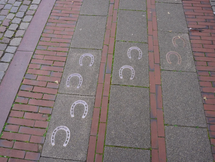 Pflasterweg mit Hufeisenmustern im Bodenbelag, flankiert von roten Backsteinabschnitten.