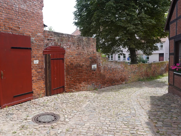 Historische Stadtmauer in Verden mit rotem Tor und Pflasterstraße, umgeben von alten Backsteingebäuden.