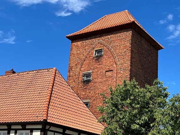 A brick tower with red bricks, with a blue sky above it; partially obscured half-timbered houses in the foreground.