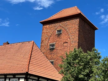 Alter Wehrturm Verden Backsteinturm mit roten Ziegeln, darüber ein blauer Himmel; teilverdeckte Fachwerkhäuser im Vordergrund.