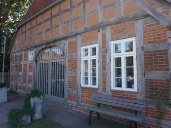 Fachwerkgebäude mit roten Backsteinen, Holzbalken und banknahe Eingangsbereich; drei weiße Fenster.
