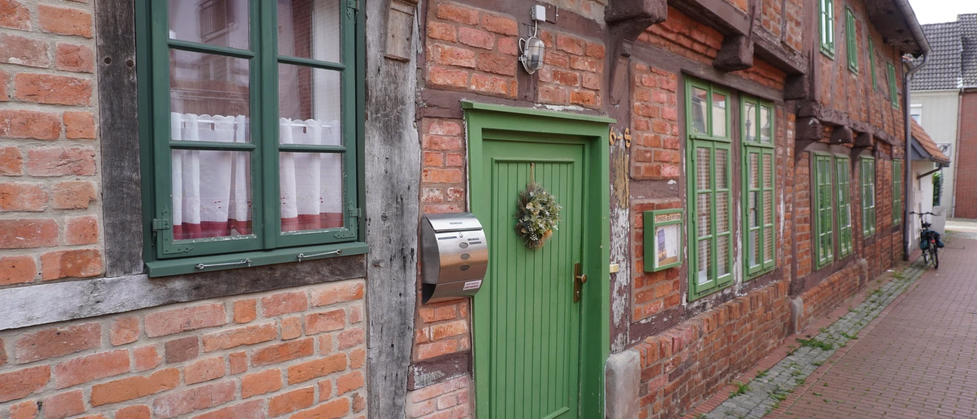 Fachwerkhäuser mit roten Ziegelwänden, weißen Fenstern und grünen Türen entlang einer Pflastergasse.