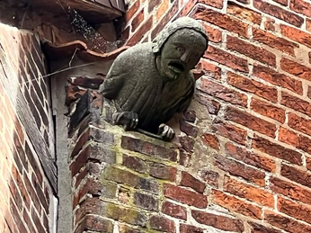 Steinerne Skulptur eines schreienden Gesichts mit langen Haaren ragt aus der Backsteinwand hervor.A stone sculpture of a screaming face with long hair protrudes from the brick wall.En stenskulptur af et skrigende ansigt med langt hår stikker ud fra murstensvæggen.Een stenen beeld van een schreeuwend gezicht met lang haar steekt uit de bakstenen muur.