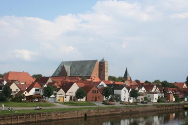 Dom Verden Eine mittelalterliche Kathedrale überragt ein Dorf mit roten Dächern am Ufer eines ruhigen Flusses.