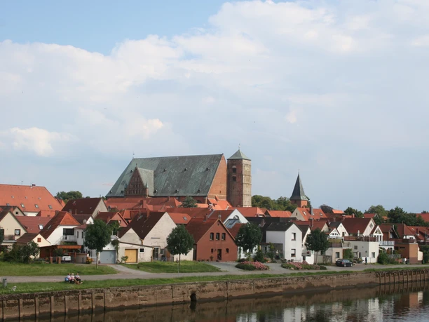 Dom Verden Eine mittelalterliche Kathedrale überragt ein Dorf mit roten Dächern am Ufer eines ruhigen Flusses.