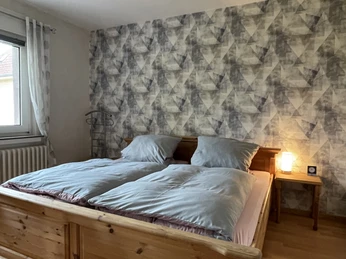 Schlafzimmer mit Doppelbett aus Holz, blauer Bettwäsche, gemusterter Tapete und kleinem Nachttisch.