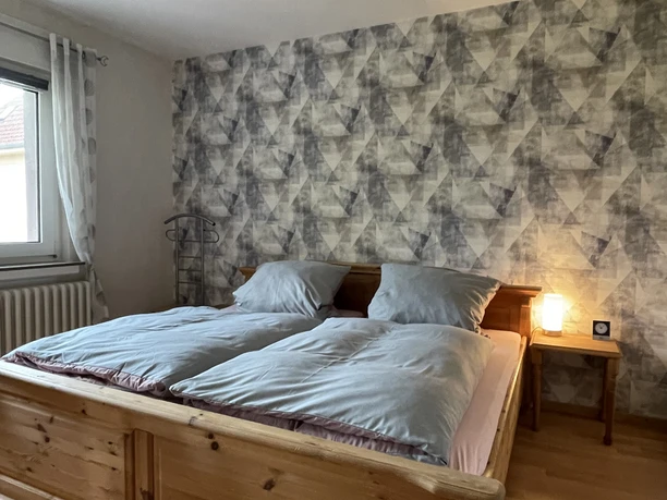 Schlafzimmer mit Doppelbett aus Holz, blauer Bettwäsche, gemusterter Tapete und kleinem Nachttisch.