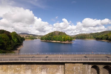 Aggertalsperre Ein Radfahrer überquert eine historische Steinbrücke am idyllischen Biggesee, umgeben von Wäldern.