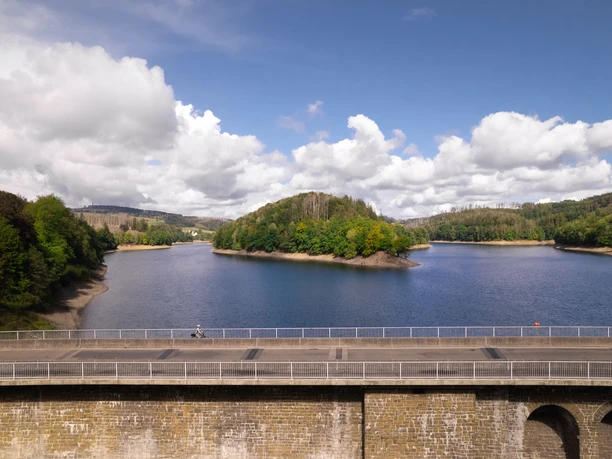 Aggertalsperre Ein Radfahrer überquert eine historische Steinbrücke am idyllischen Biggesee, umgeben von Wäldern.