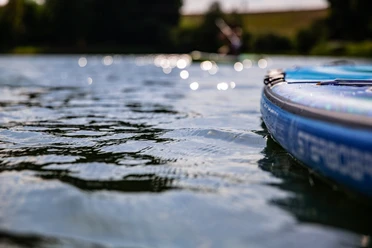 Stand-up-Paddling Nahaufnahme eines blauen SUP-Boards auf einem ruhigen See mit glitzerndem Wasser und unscharfen Ufern.