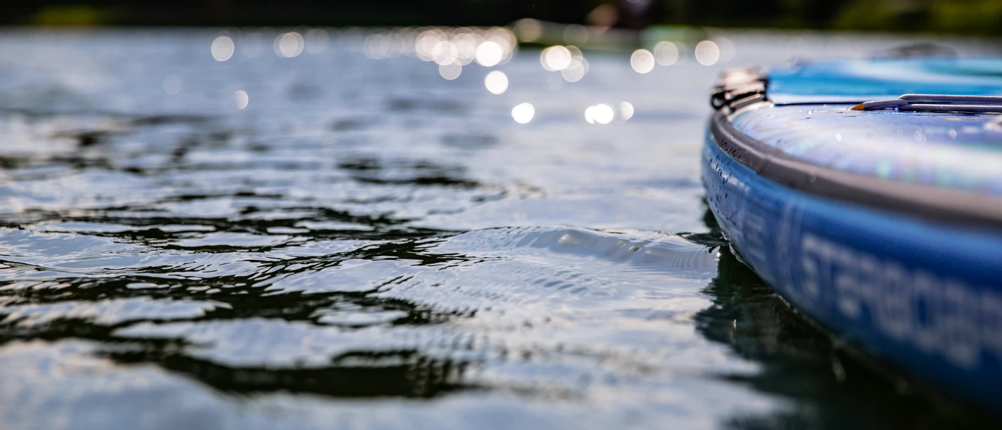 Stand-up-Paddling Nahaufnahme eines blauen SUP-Boards auf einem ruhigen See mit glitzerndem Wasser und unscharfen Ufern.