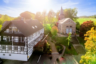 Blick auf Kürten-Delling Fachwerkhaus und historische Kirche bei Sonnenschein, umgeben von Bäumen und grüner Landschaft.