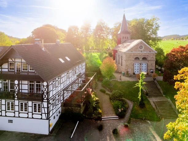 Blick auf Kürten-Delling Fachwerkhaus und historische Kirche bei Sonnenschein, umgeben von Bäumen und grüner Landschaft.